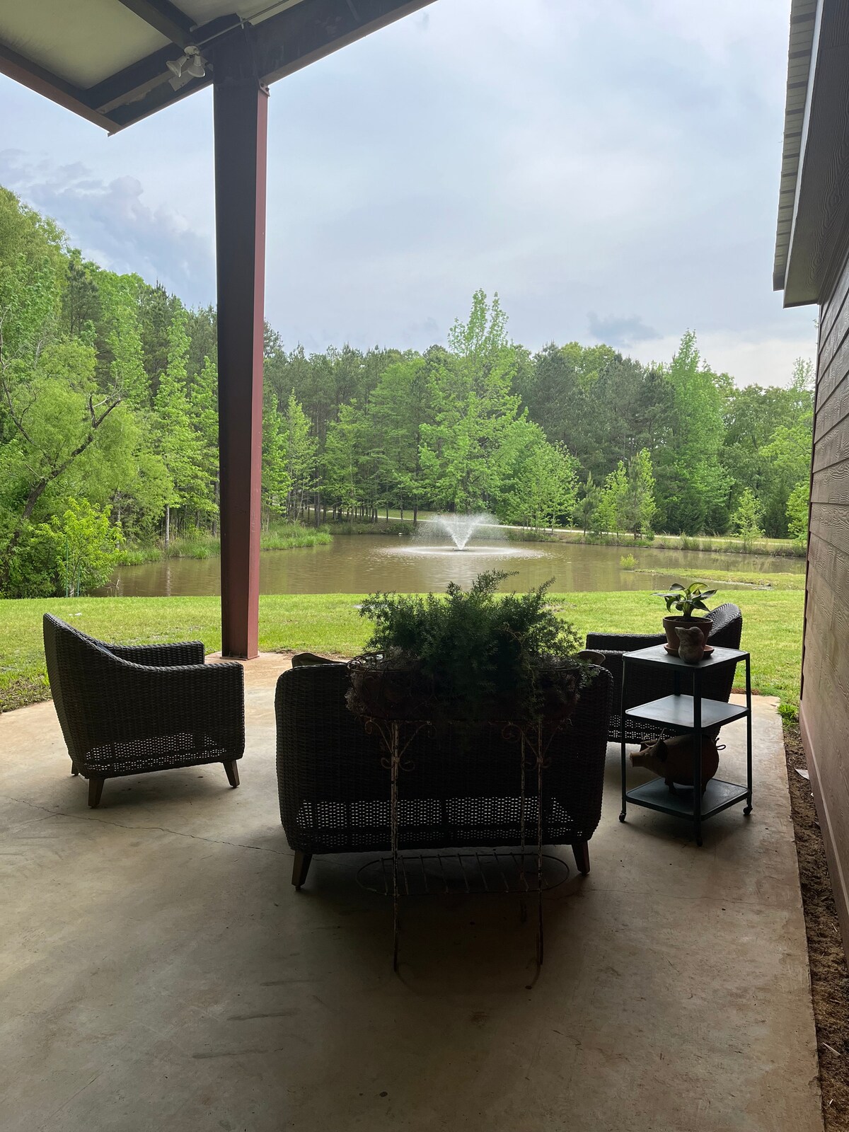 A covered outdoor seating area includes four wicker chairs arranged around a small table, providing a view of a serene pond with a fountain. Lush greenery surrounds the area, enhancing the peaceful ambiance.