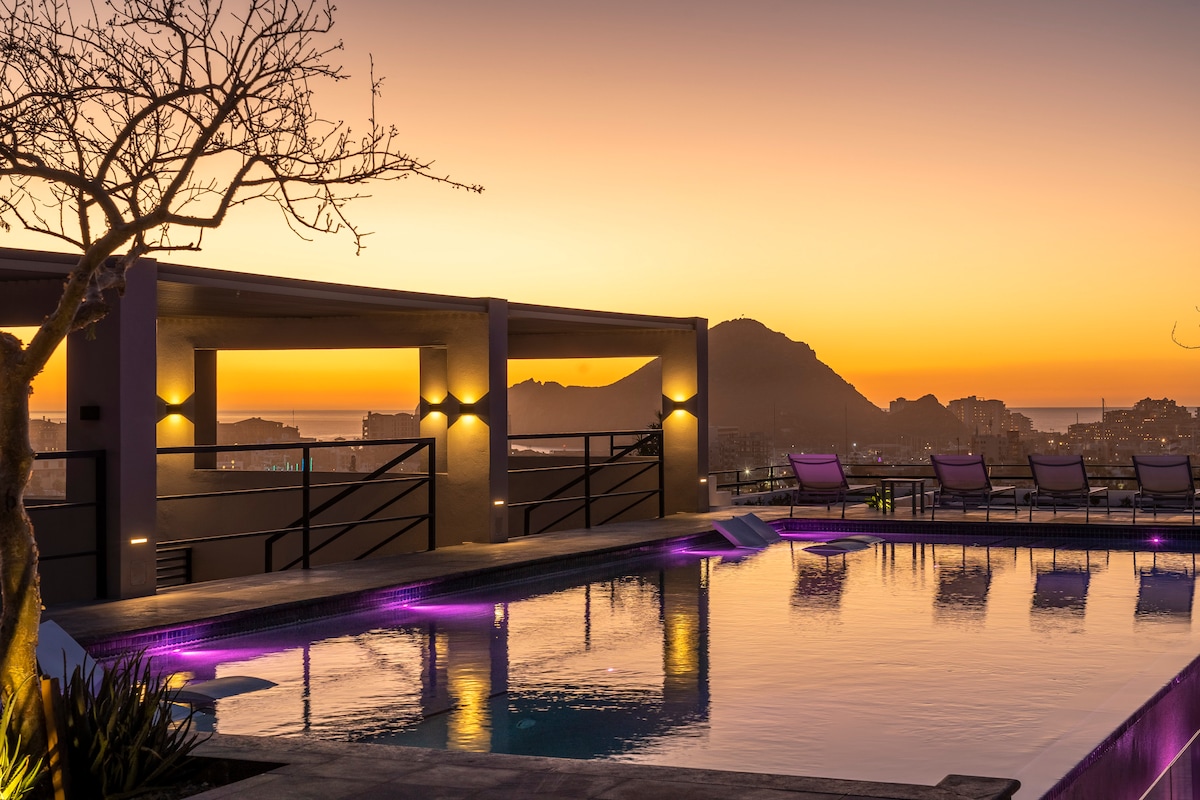 A serene rooftop pool reflects a vibrant sunset, surrounded by lounge chairs arranged along the edge. Soft purple lighting enhances the tranquil atmosphere, while distant hills and city lights create a picturesque horizon.