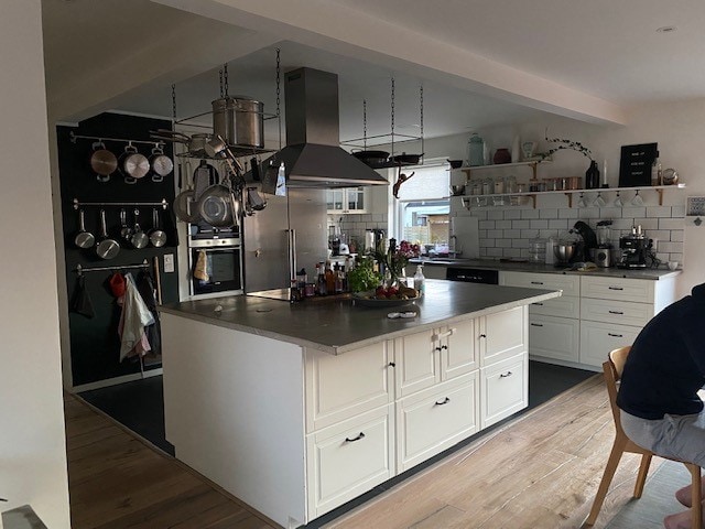 An inviting kitchen features a spacious island with a gray countertop and ample storage beneath. Cooking utensils are neatly organized above and behind, while modern appliances are integrated seamlessly into the cabinetry. Natural light fills the room, creating a warm and functional space.