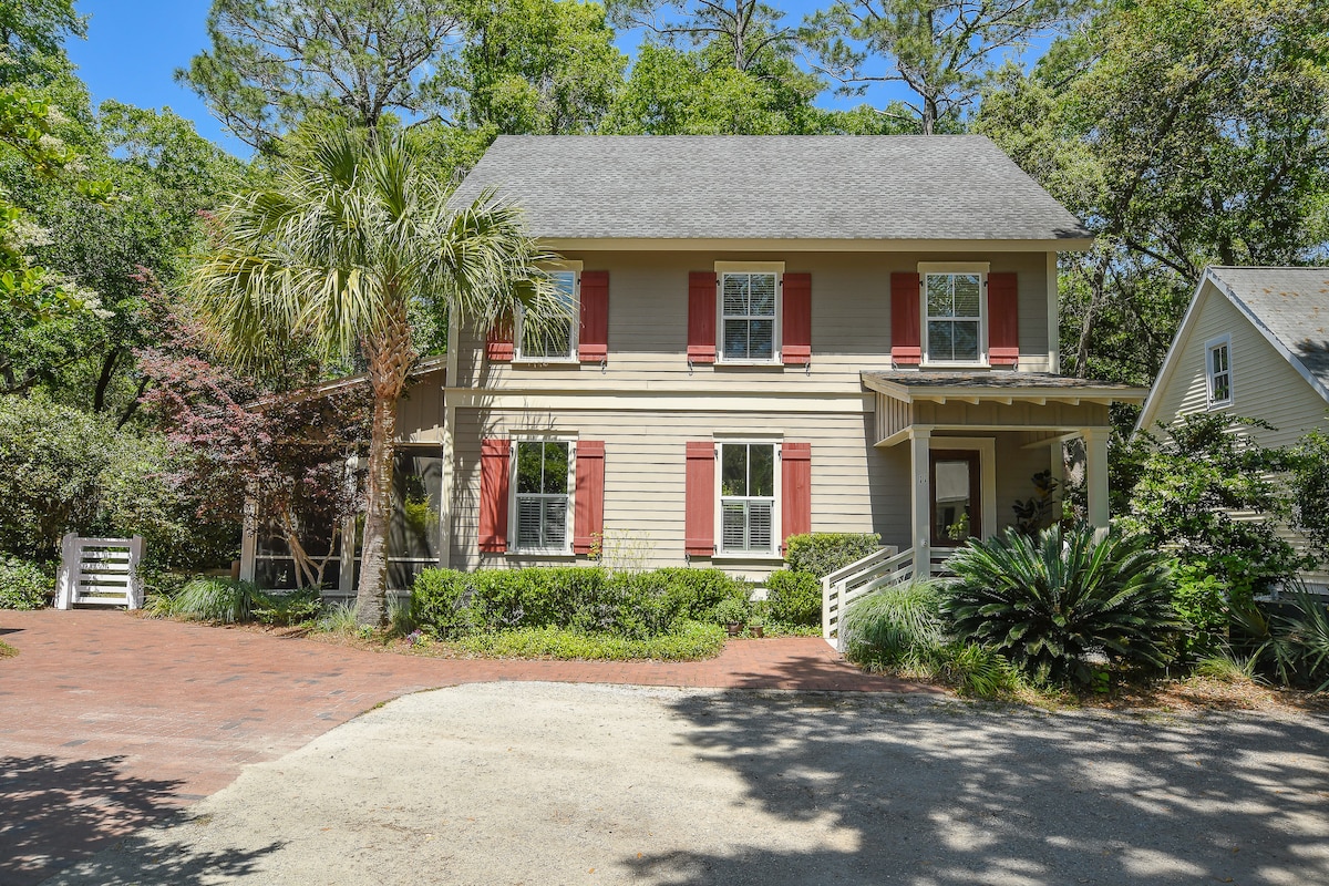 A two-story house with light yellow siding and red shutters is surrounded by lush greenery. A brick pathway leads to the welcoming front porch, framed by mature trees and manicured shrubs. The private driveway features a compact gravel area for parking.