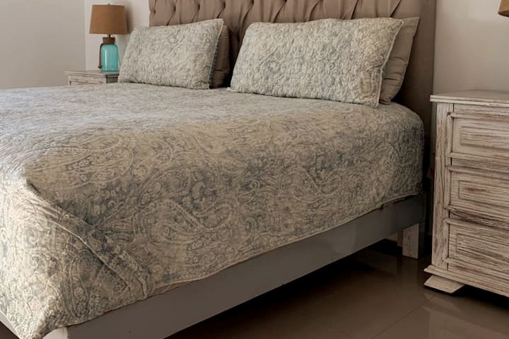 Dhoma kryesore e gjumit ka një krevat të madh dhe të bardhë luksoz, si dhe detaje dhe ngjyra që krijojnë një atmosferë plazhi.