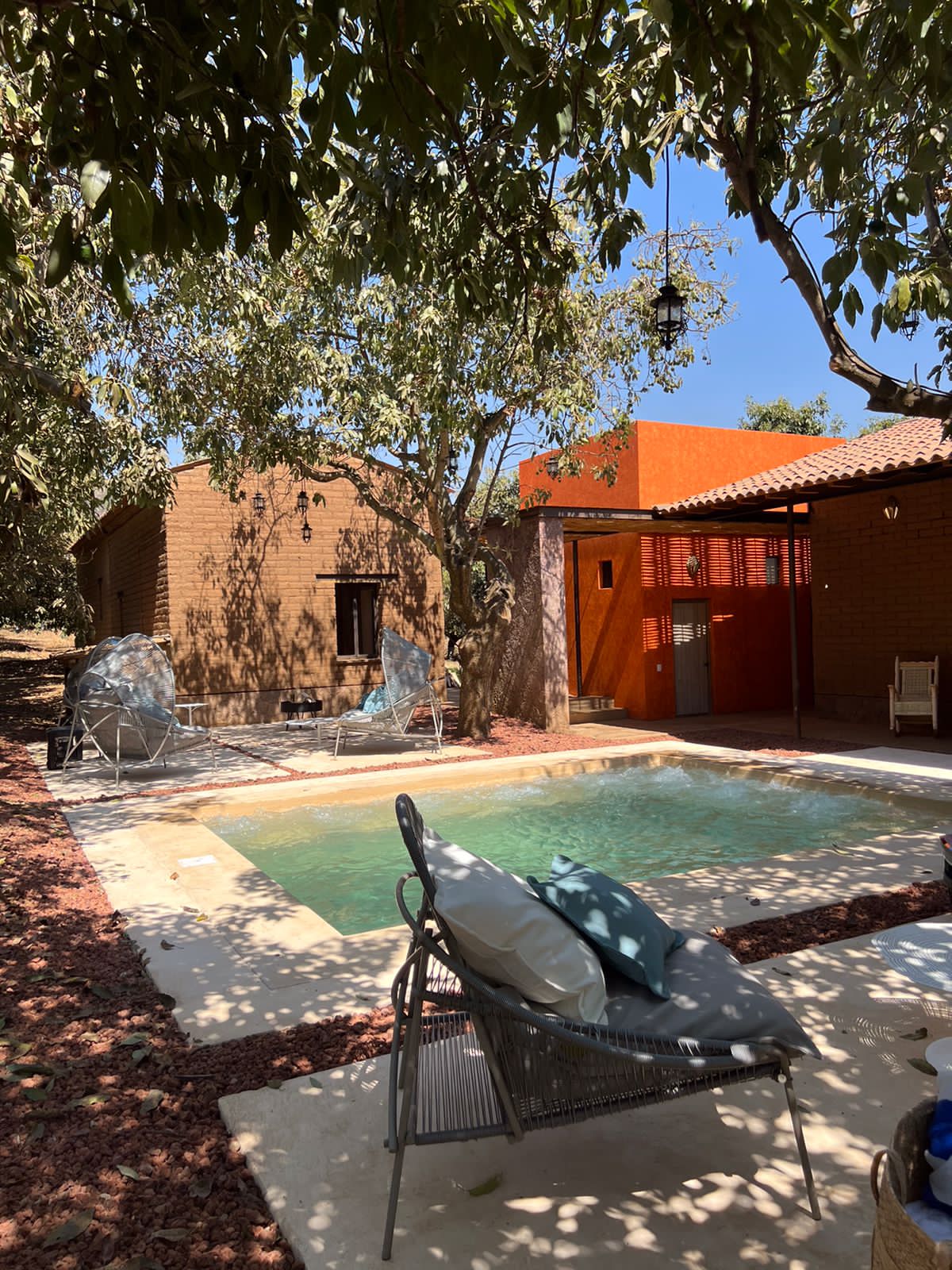 A tranquil outdoor area features a pool surrounded by shaded foliage. A comfortable seating arrangement nearby showcases plush pillows. The rustic adobe structure in the background complements the natural setting with warm tones and terracotta accents.