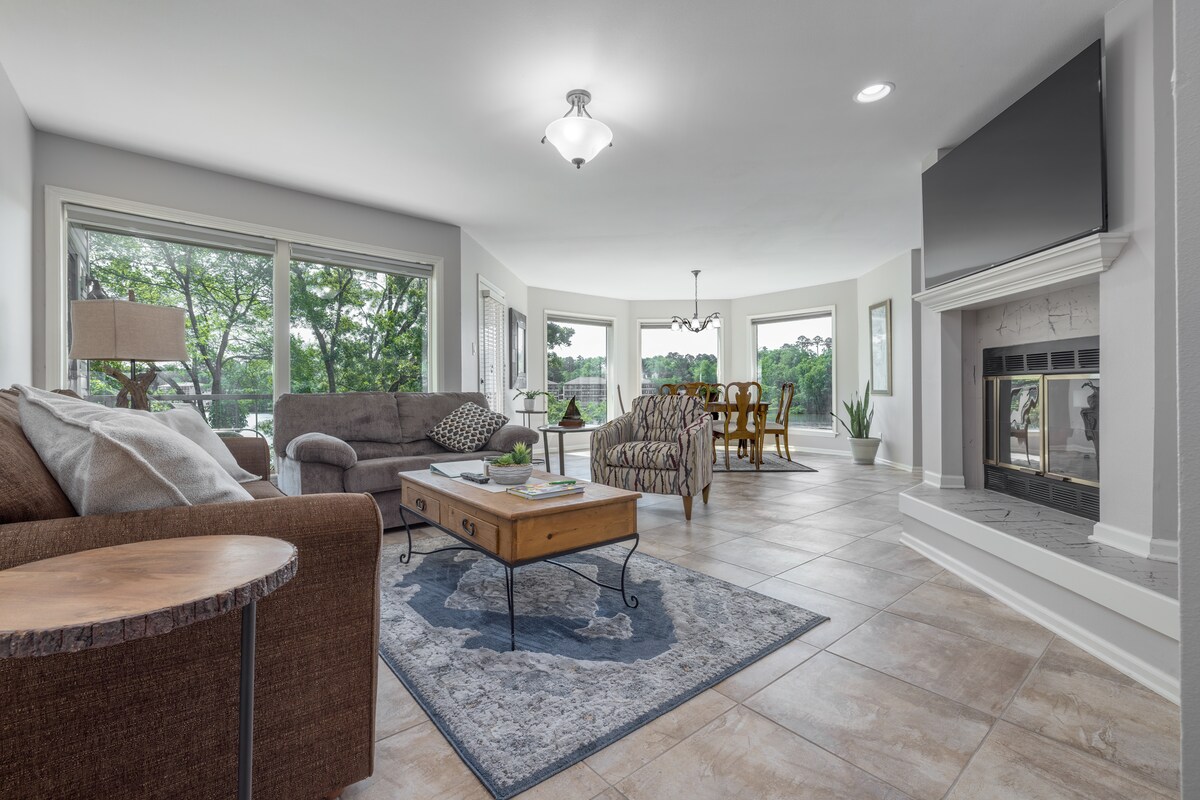 A spacious living area features large windows allowing natural light to fill the space. A comfortable sofa and armchair are arranged around a central coffee table, with a decorative rug underneath. A dining table with multiple chairs is visible in the background, and a fireplace adds a focal point.