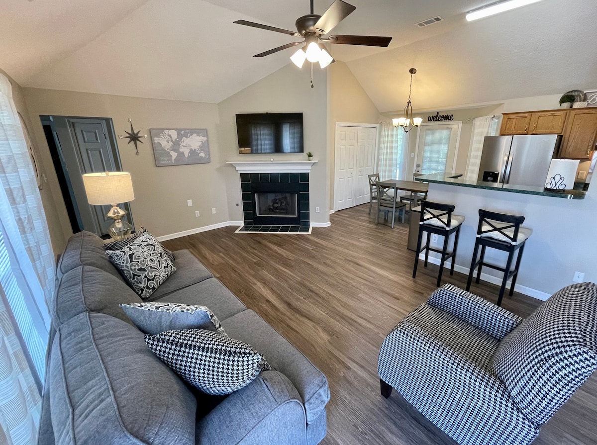 An inviting living space features a gray sofa with patterned cushions, complemented by a cozy armchair. A modern fireplace is positioned beneath a wall-mounted TV, while a dining area with bar stools can be seen in the background. Natural light enhances the warm atmosphere.
