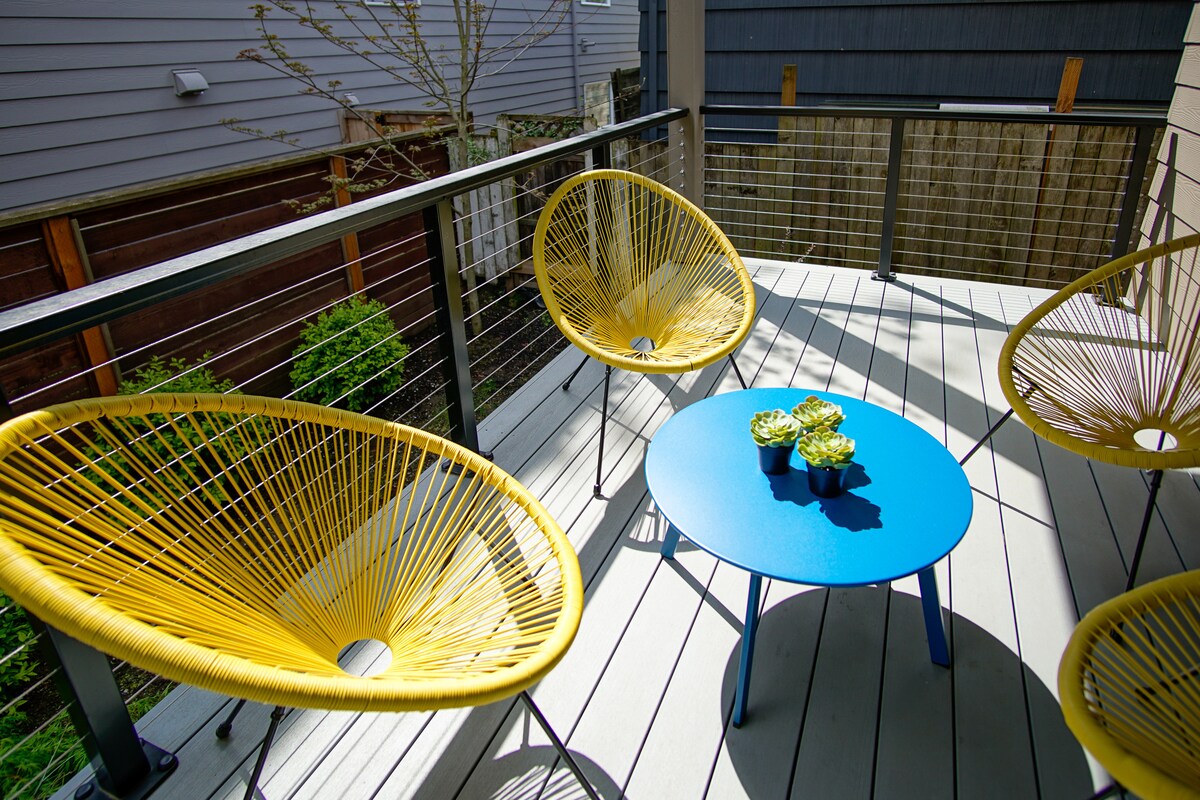 A serene outdoor seating area is presented featuring three vibrant yellow chairs surrounding a blue table. Potted plants add a touch of greenery, complementing the peaceful surroundings. The deck is constructed of light-colored wood, providing a refreshing outdoor space.