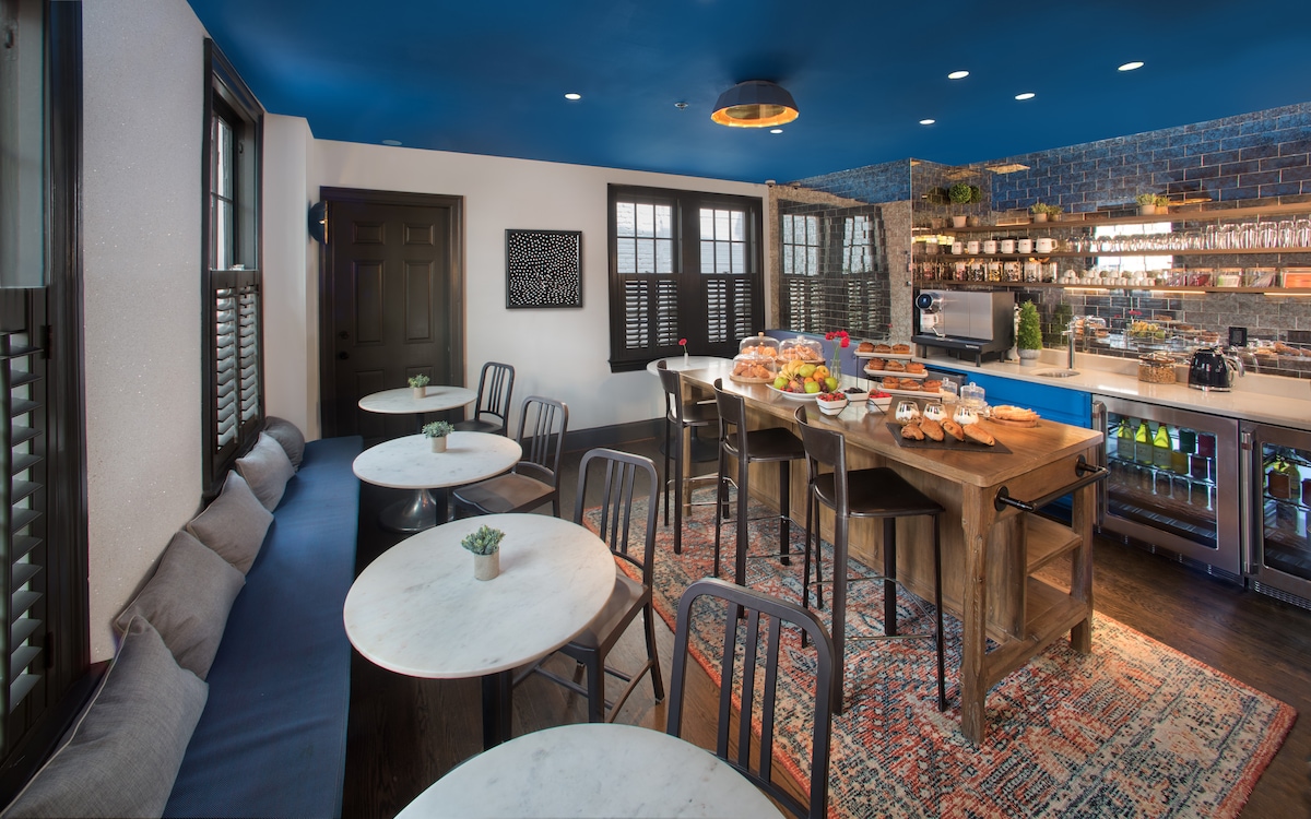 A cozy communal dining area features multiple round tables with marble tops and sturdy black chairs. A wooden counter showcases a selection of breakfast items. Large windows allow natural light to fill the space, highlighting the tasteful decor and inviting atmosphere.