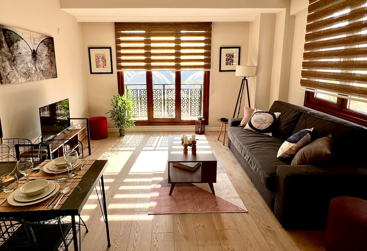 A living area features a comfortable dark sofa adorned with various cushions, complemented by a table set for dining. A television is positioned nearby, and plants add a touch of greenery. Natural light filters through stylish wooden blinds, creating a warm environment.