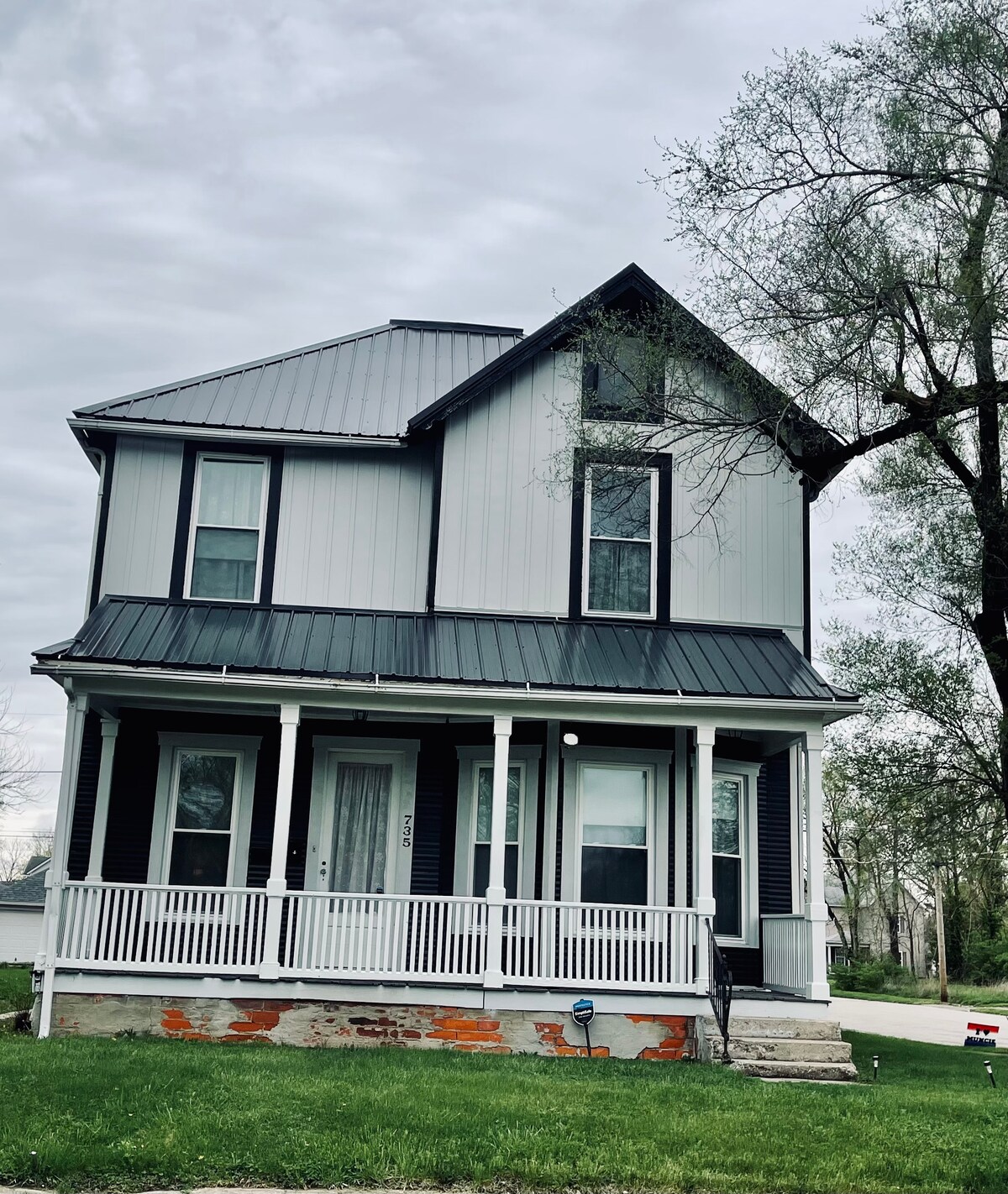 The exterior of a two-story house is featured, showcasing a light gray facade with dark trim. A porch with white railings extends across the front, providing access to the main entrance. Large windows offer natural light, and surrounding greenery is visible in the yard.