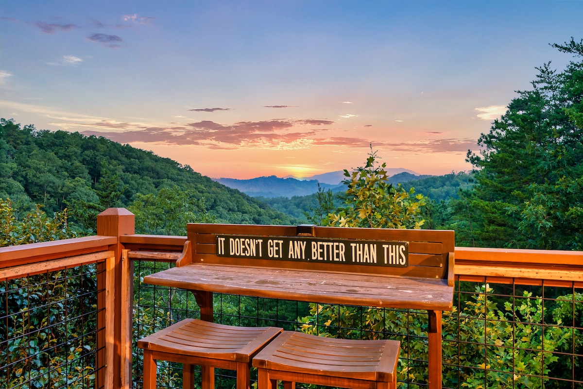 A scenic outdoor seating area features a wooden table and two stools, overlooking a vast mountainous landscape. The horizon displays soft pastel colors of sunset, with lush greenery abundant in the foreground and hills in the distance.