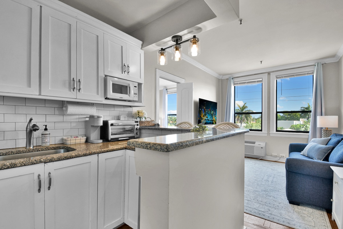 The kitchenette area is equipped with white cabinetry and a granite countertop. An oven, microwave, and sink are visible, along with ample storage space. A living area features a blue sofa and a large TV, with sunlight coming through two windows showcasing views of palm trees.