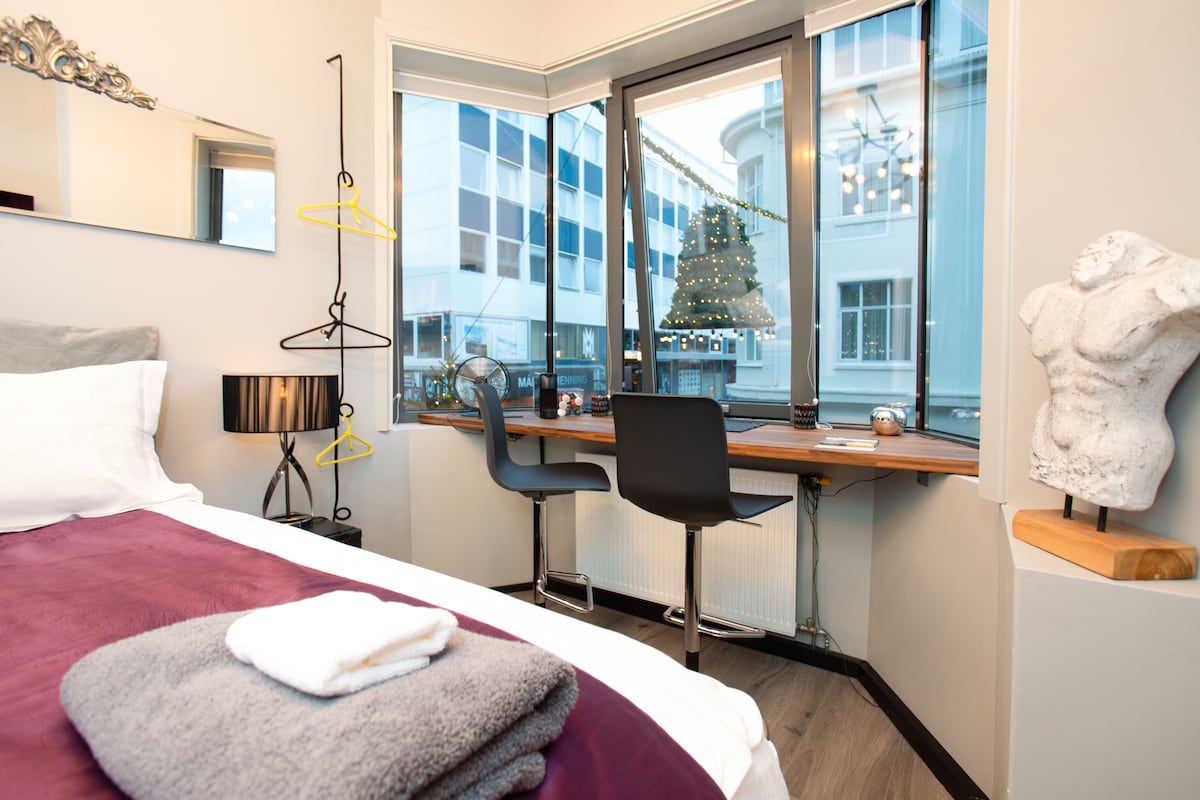 A cozy corner of the studio features a bay window with a view over the Main Street. A small desk with a chair is positioned next to the window, complemented by a stylish lamp. A frosted towel is neatly folded on the bed, with a decorative sculpture nearby.