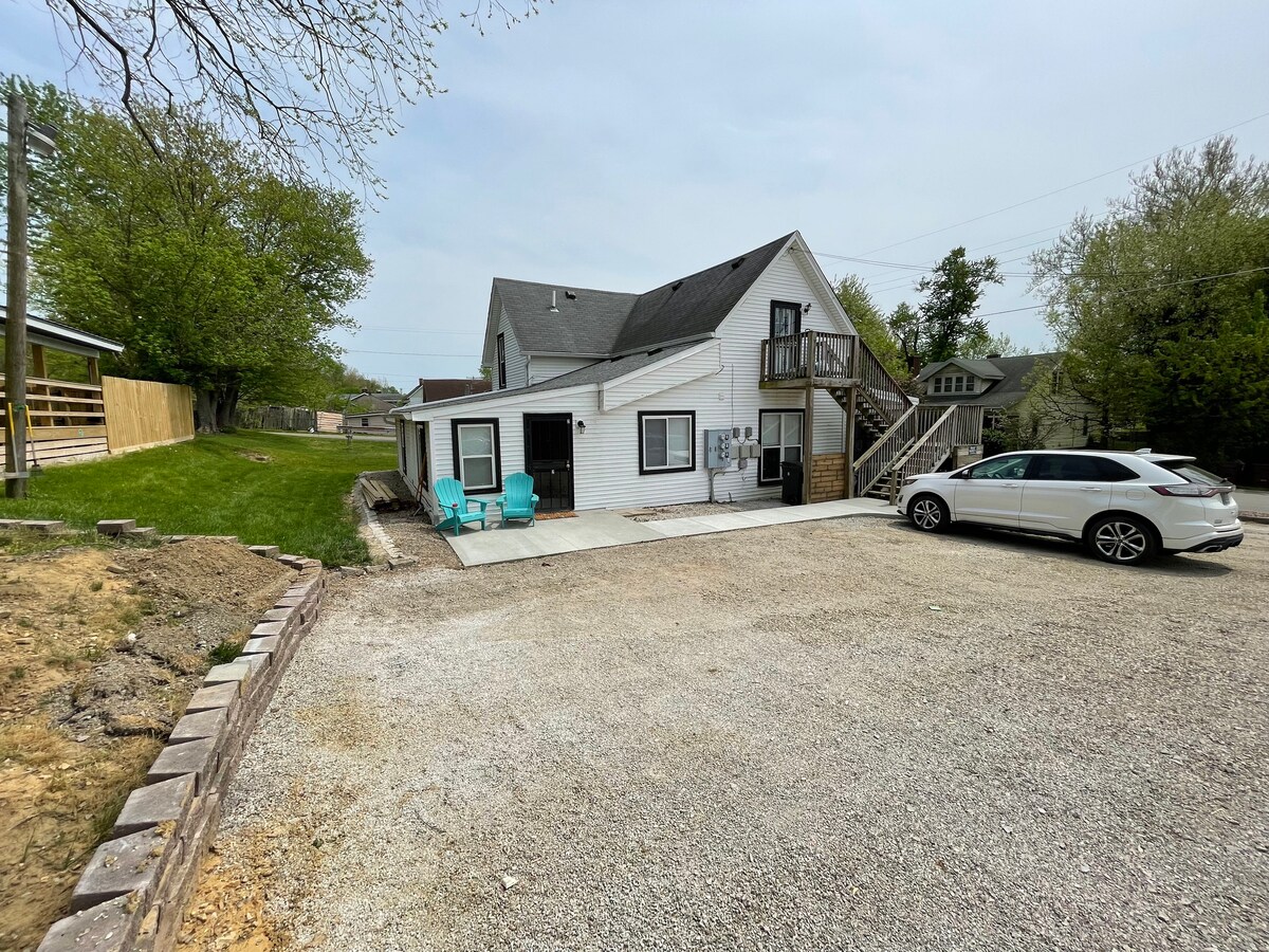 The exterior view displays a two-story triplex with a white facade and a staircase leading to the upper level. Two turquoise chairs are positioned on the patio area, and a white vehicle is parked in the gravel driveway. Lush greenery surrounds the property.