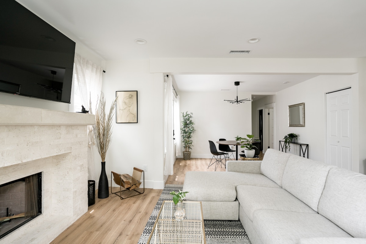 A spacious living area features a light-colored sectional sofa positioned near a stylish fireplace. A flat-screen TV is mounted on the wall, and natural light is invited through sheer curtains. A dining area is visible in the background, showcasing a modern table and chairs.