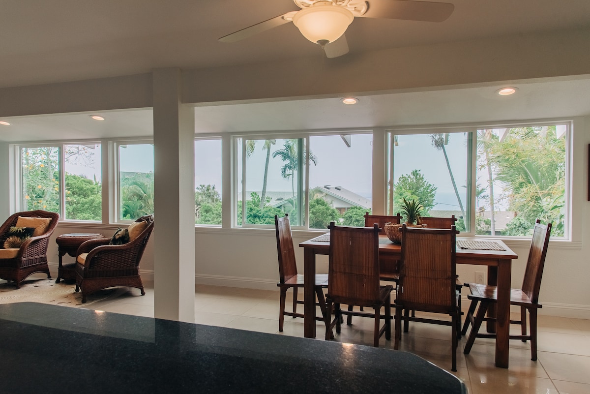 A bright dining area presents a wooden table surrounded by chairs, with large windows offering views of palm trees and the ocean. Comfortable wicker seating is positioned nearby, allowing for relaxation while enjoying the scenic surroundings.