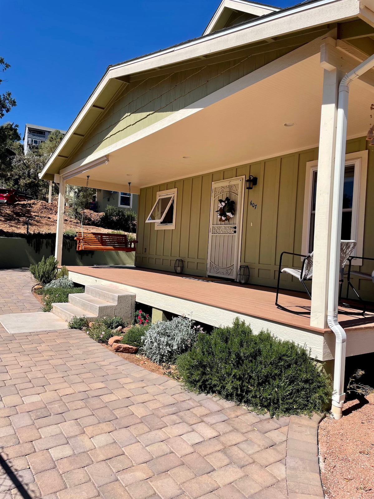 A charming front porch features a wooden swing and a chair, set against a light green cottage exterior. The pathway is composed of pavers leading to steps adorned with a welcoming mat. Green shrubs and desert plants create a natural border around the porch.