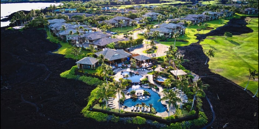 An aerial view captures the expansive Hali'i Kai Resort, showcasing a beautifully landscaped area with lush greenery and multiple structures. The central pool area is surrounded by lounge chairs and palm trees, offering a refreshing retreat amidst volcanic rock formations.