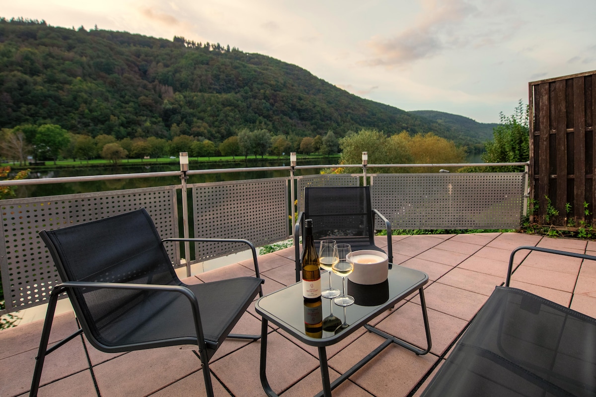 A terrace features modern outdoor furniture, including two chairs and a glass table. A bottle of wine and two glasses sit ready for enjoyment. The background showcases a scenic view of trees and hills along the Mosel River, creating a tranquil setting.