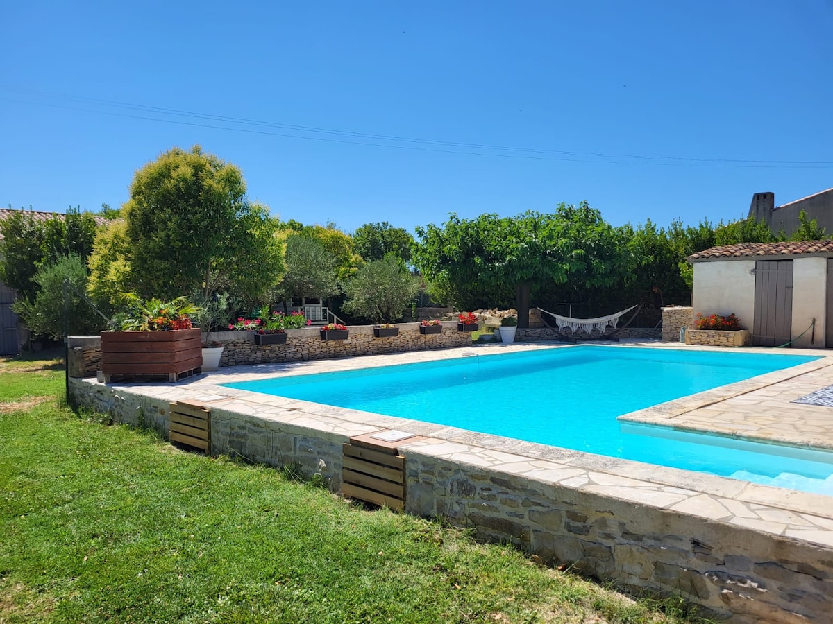 A clear blue swimming pool is framed by a stone patio and surrounded by lush greenery, including trees and flowering plants. A hammock is visible nearby, adding to the tranquil outdoor ambiance of the space.