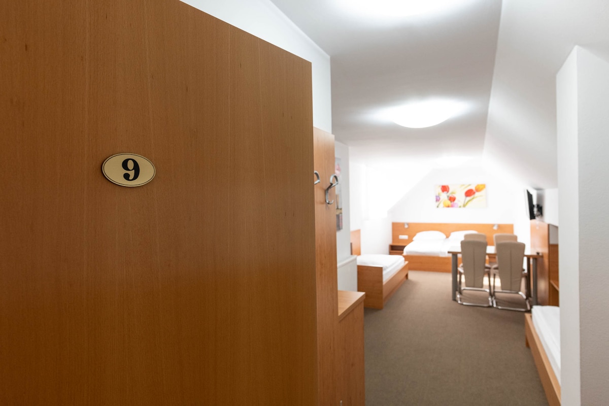 A spacious room is accessible through a wooden door marked with the number 9. The room features light-colored walls and a neutral carpet. Two beds are positioned alongside a wooden dining table and chairs. A television is mounted on the wall, and soft lighting enhances the interior.