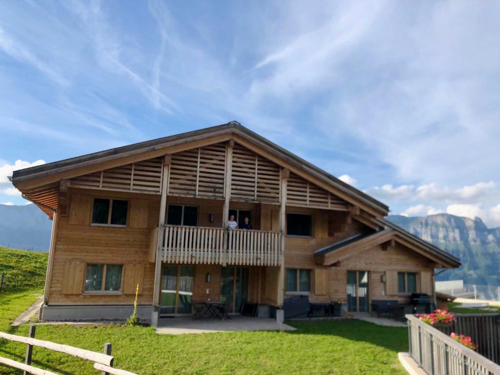 A chalet-style building with wooden exterior is set against a backdrop of rolling green hills and mountains. A balcony extends from the second floor, providing outdoor seating options. Large windows enhance natural light, creating a welcoming appearance in a serene landscape.