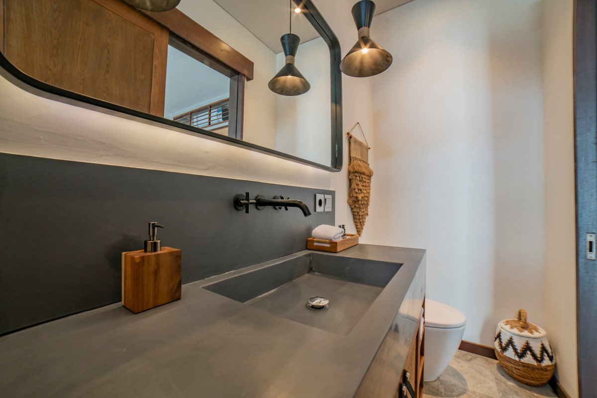 A modern bathroom features a sleek concrete sink with a minimalist faucet, complemented by ambient lighting from pendant fixtures. A large mirror reflects the design elements, while natural materials accentuate the space. A simple toilet is positioned to the side, emphasizing the uncluttered layout.