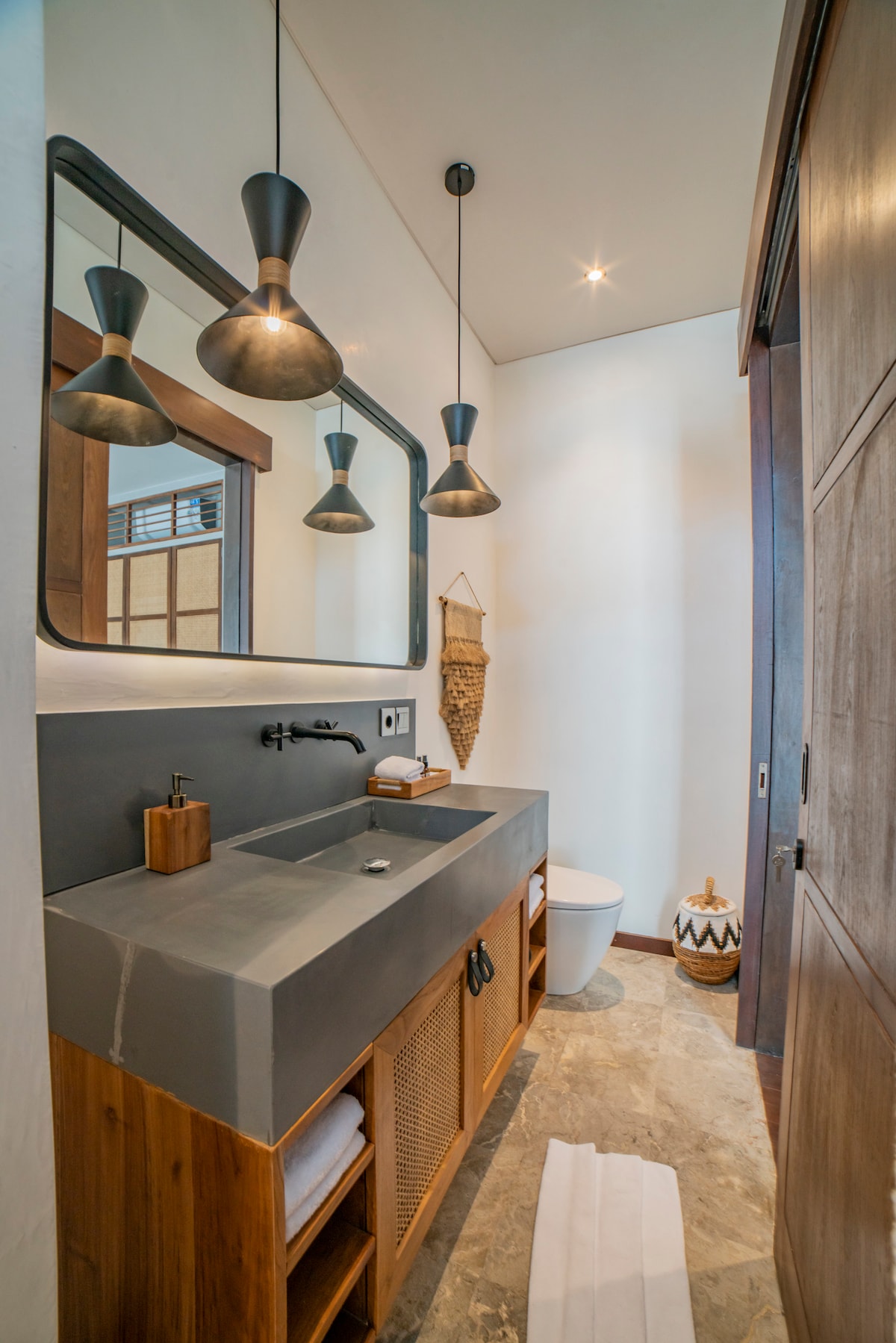A modern bathroom features a wooden vanity with a wide sink, complemented by two pendant lights above. A large mirror reflects the space, while soft white towels are neatly arranged. The room is illuminated by natural light, showcasing stone flooring and minimalist decor.
