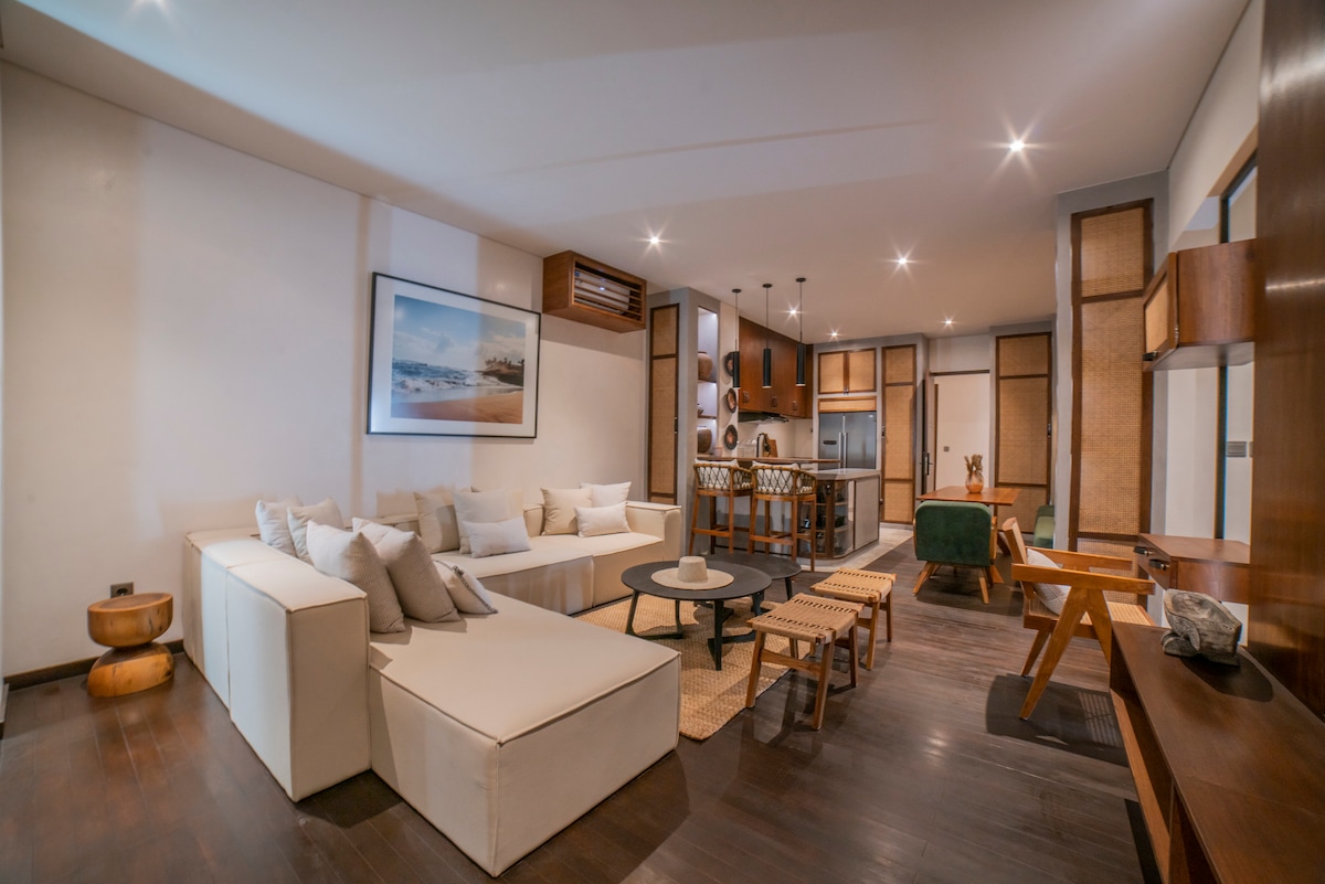 The spacious living area features a light-colored sectional sofa arranged around a circular table. Decorative cushions add comfort, while a modern kitchen is visible in the background. Warm wooden accents and ambient lighting create a welcoming environment.