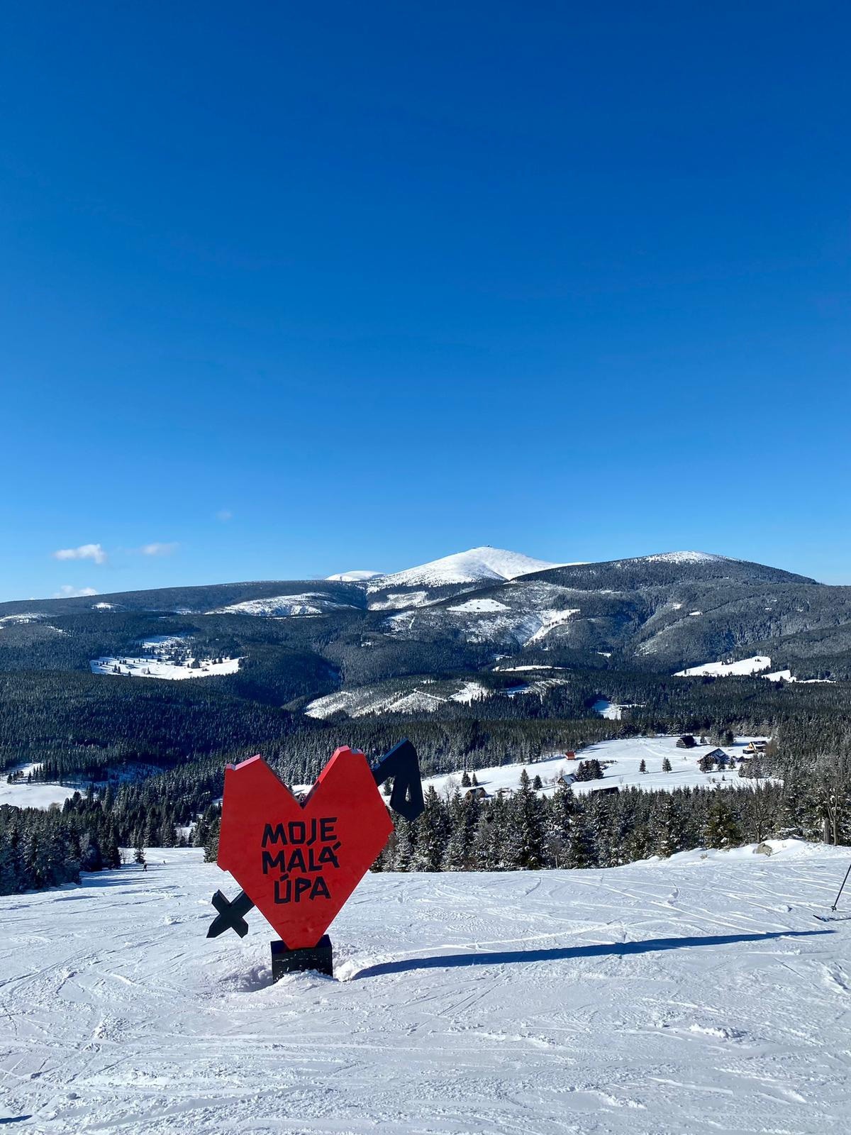 A vibrant red heart sculpture stands prominently with the words 'MOJE MALA ÚPA.' Snow-covered slopes extend into the background, leading to a mountainous landscape under a clear blue sky. The scene conveys a serene winter atmosphere.