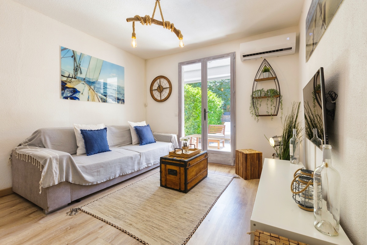 A light-filled living area features a comfortable sofa with blue cushions and a decorative throw. A wooden coffee table and a minimalist TV stand are arranged around a soft area rug. Natural light streams in from large glass doors that lead to a private terrace.