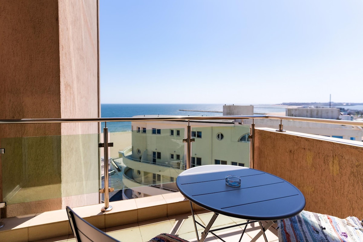 A balcony presents an inviting outdoor space, featuring a round table accompanied by two chairs. The view includes expansive ocean waters and a clear blue sky, creating a serene backdrop for relaxation.