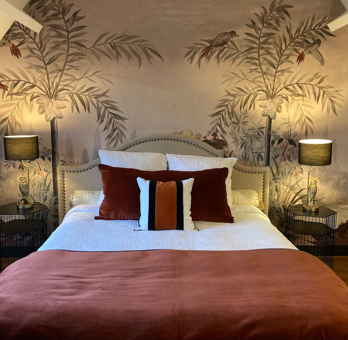 A cozy sleeping area features a spacious bed adorned with a white duvet and earthy-toned pillows. The backdrop showcases a mural of tropical plants, while bedside tables with lamps provide soft lighting, enhancing the room's inviting ambiance.