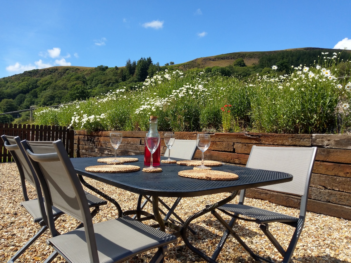 An outdoor dining area is set with a round table and six metal chairs, arranged on a gravel surface. A bottle of sparkling drink and glasses are positioned at the center. Lush greenery and wildflowers can be seen in the background against a clear blue sky.