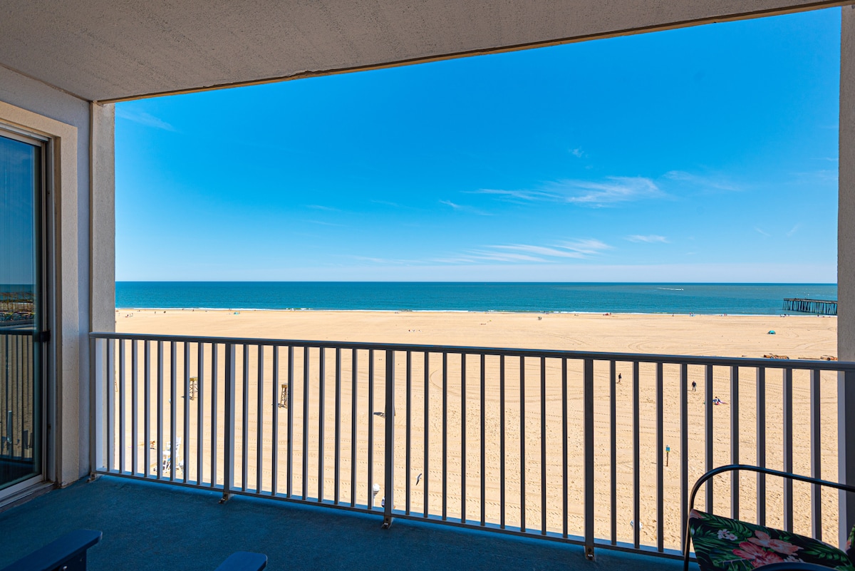 The balcony offers a wide view of the sandy beach and ocean, complemented by a clear blue sky. A railing frames the scene, while the horizon meets the tranquil water. The space is designed for relaxation with seating options positioned to enjoy the view.