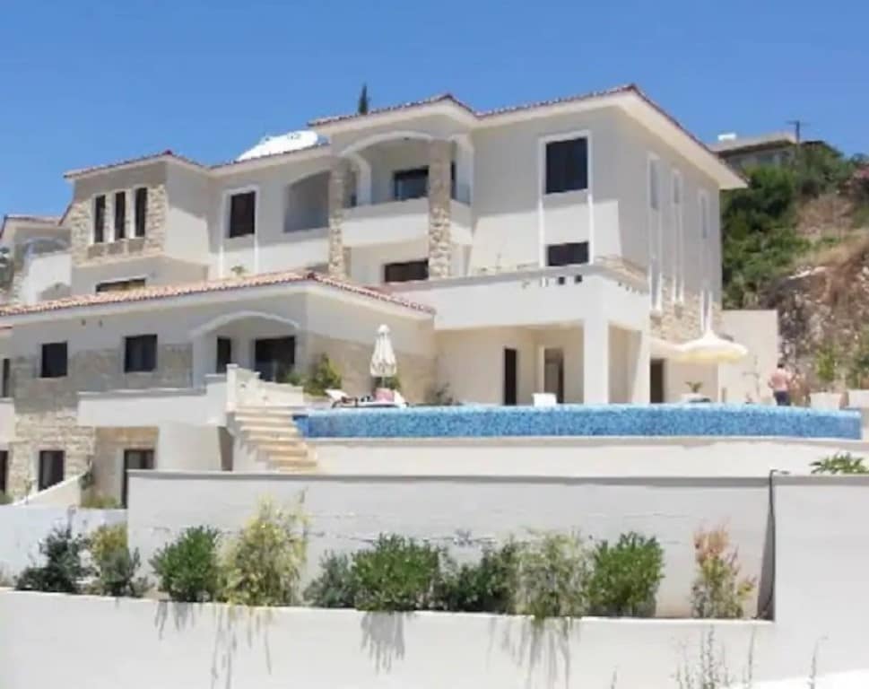 The stunning exterior of a modern villa is highlighted, featuring a multi-level structure with stone accents. A private infinity pool is situated on a terrace, surrounded by lush greenery. Large windows allow for ample light, creating an inviting atmosphere. A covered area is visible for outdoor relaxation.