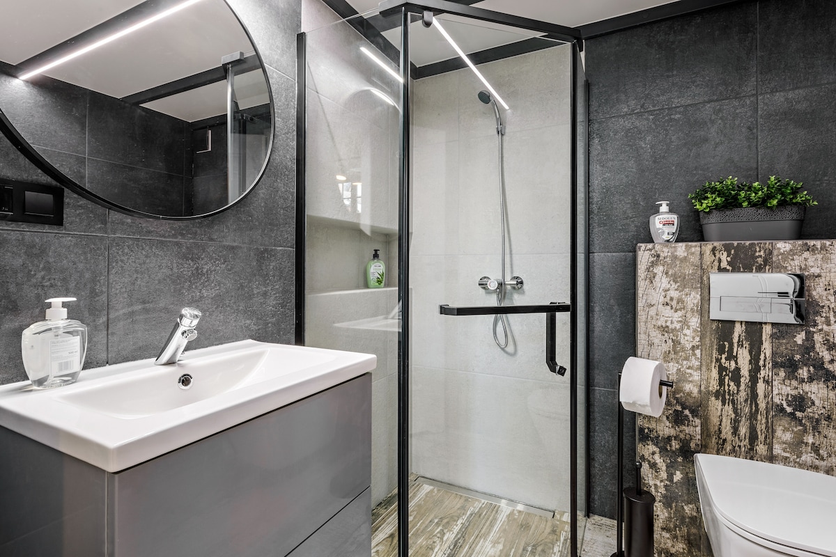 A modern bathroom features a sleek, gray vanity with a round mirror above. A glass-enclosed shower is located in one corner, showcasing clean lines and contemporary fixtures. A potted plant adds a touch of greenery, while textured tiles in neutral tones complement the overall design.