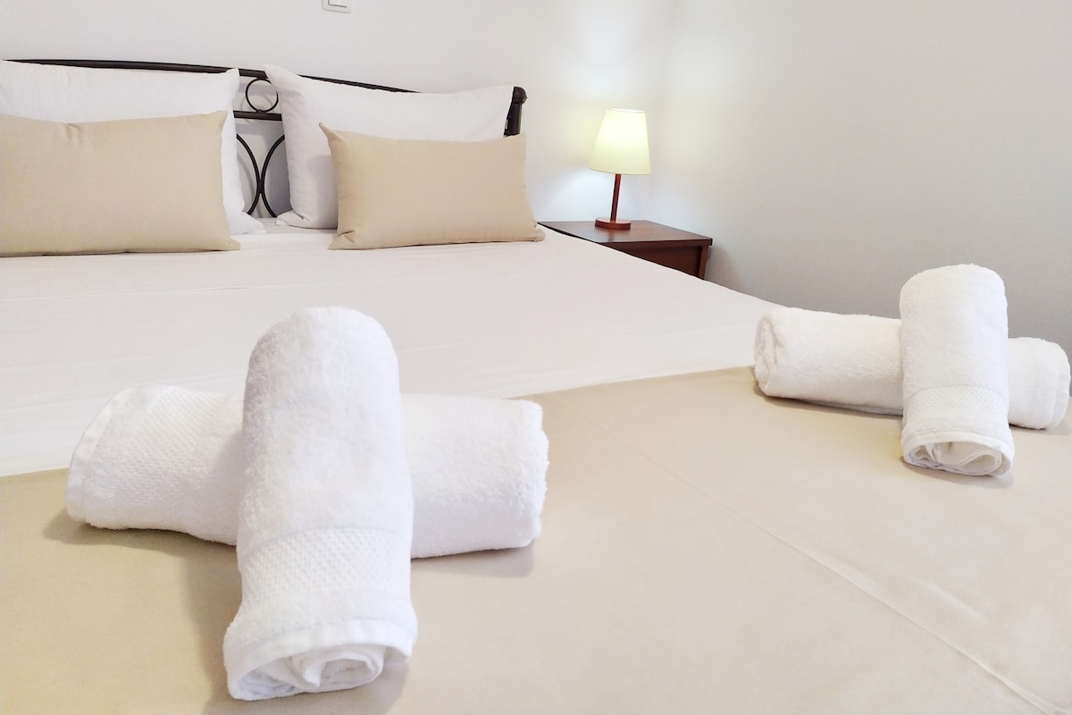 A spacious bed is dressed in crisp white linens, complemented by soft beige accents. Rolled white towels are neatly arranged at the foot of the bed. A bedside lamp casts a gentle light, creating a calming atmosphere.