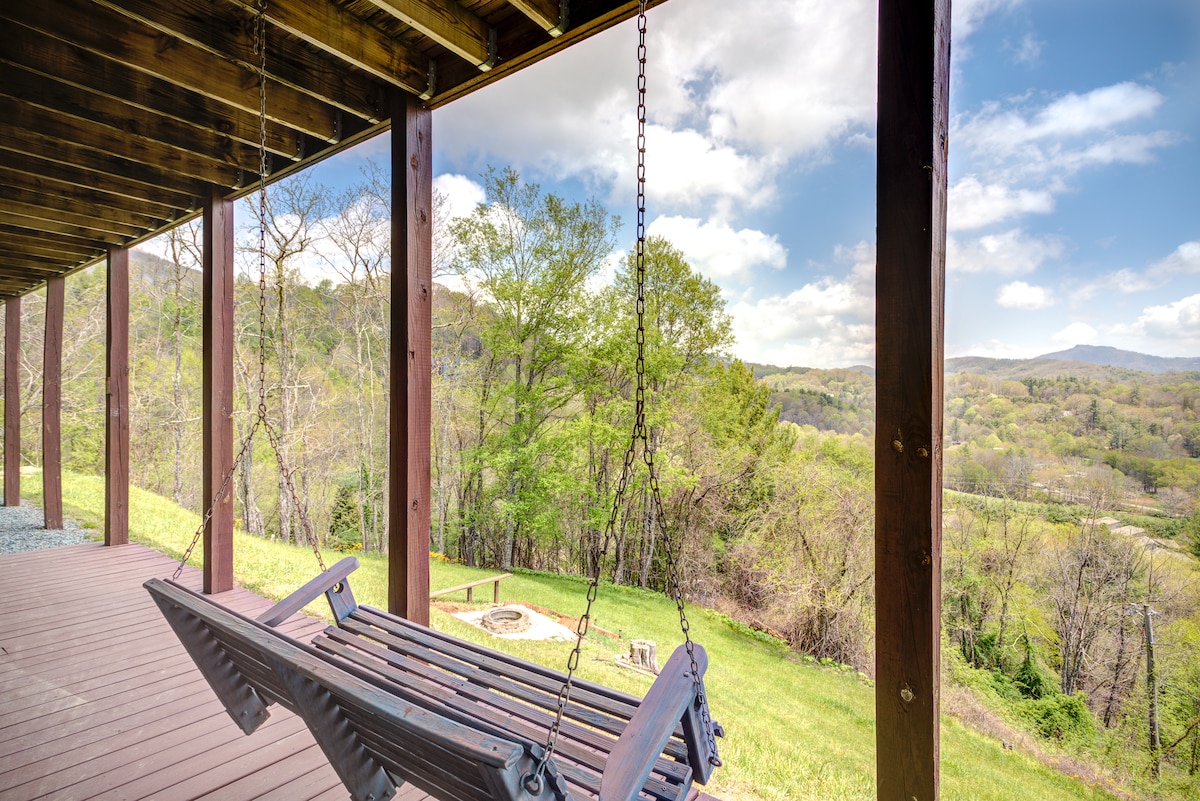 A porch swing hangs from a wooden awning, overlooking a scenic landscape filled with lush greenery. The background features rolling hills and distant mountains under a partly cloudy sky, with a fire pit visible on the grassy area below.