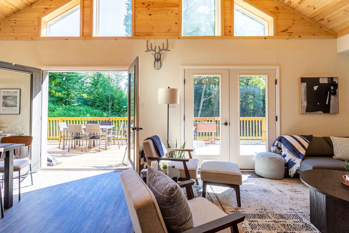 A bright living area features wooden beams and large windows allowing natural light to flood the space. Double French doors open to a deck, connecting indoor and outdoor areas. Comfortable furnishings include a plush sofa and accent chairs, enhancing the inviting atmosphere.