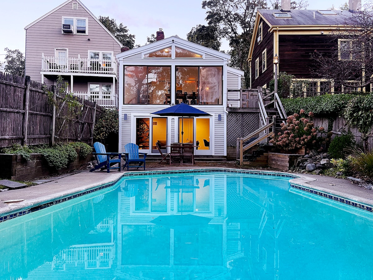 The inviting backyard features a clear pool reflecting a serene atmosphere. A patio with seating is visible near the pool. The house, with large windows, provides a bright indoor space that overlooks the outdoor area, surrounded by lush greenery and charming landscaping.