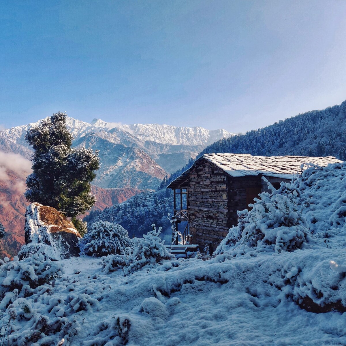 A stone cottage is partially covered in snow, surrounded by trees and shrubbery. The distant mountains are blanketed in white, contrasting with the blue sky. The serene landscape offers a glimpse of winter’s beauty in this remote location.