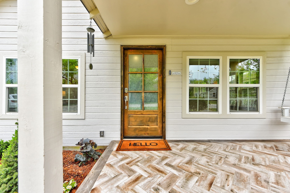 The entrance features a welcoming wooden door with a golden handle, complemented by a doormat that reads 'HELLO.' Large windows flank the door, providing natural light, while a porch swing adds a cozy touch to the space.