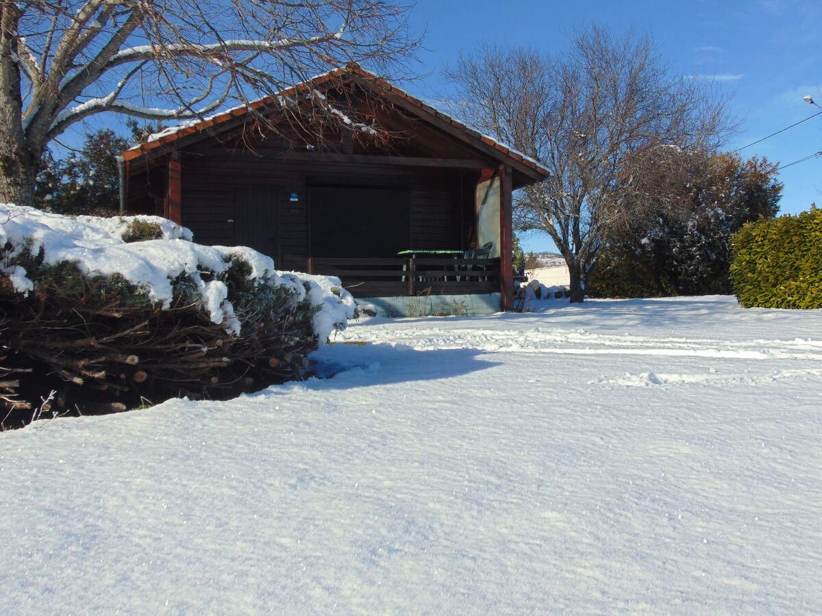 A charming chalet is surrounded by a blanket of fresh snow, with a wooden porch visible in front. A stack of firewood is neatly arranged to the left, and tall trees provide a backdrop against the clear blue sky.