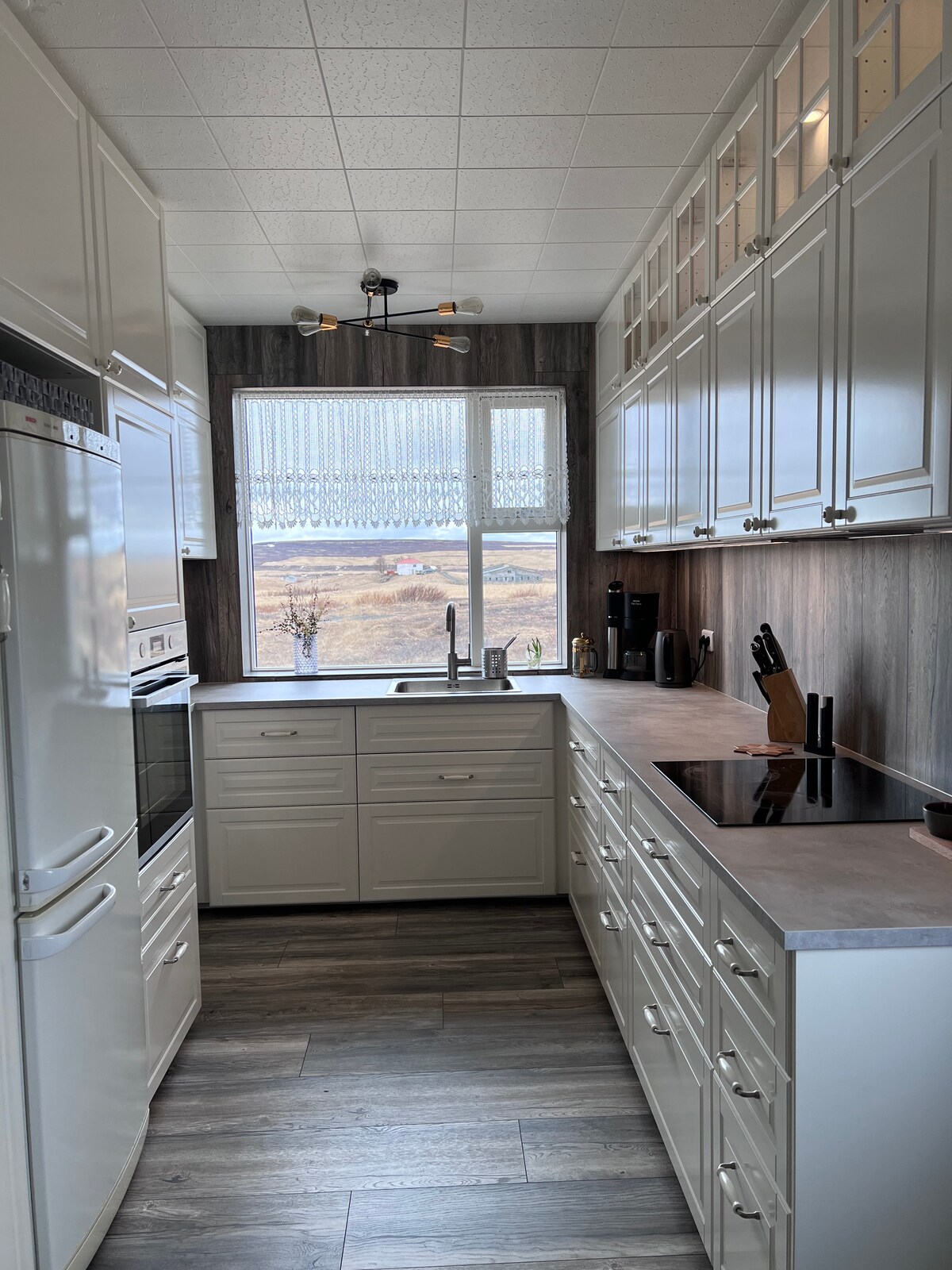 A modern kitchen features white cabinetry and a dark wood accent wall. A generously sized window allows natural light to illuminate the space, revealing scenic views of the surroundings. Countertops are equipped with essential appliances, and kitchen tools are neatly arranged for convenience.