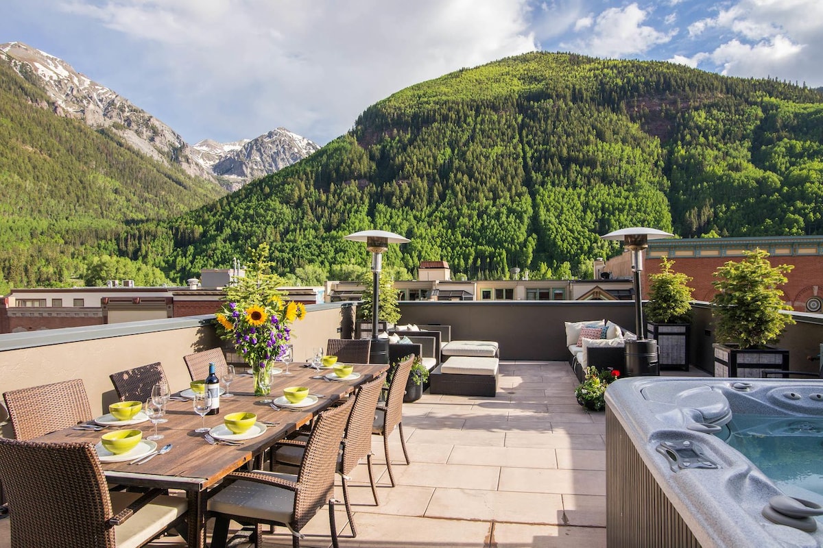 An expansive south-facing deck showcases a long dining table adorned with colorful tableware and floral arrangements. Comfortable seating areas and a hot tub are highlighted, with lush green mountains serving as a scenic backdrop. Sunlight brightens the space, enhancing the inviting atmosphere.