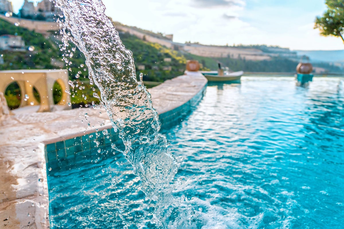 An outdoor pool is shown with clear blue water, featuring a gentle waterfall flowing into the pool. Scenic hills provide a backdrop, highlighting the lush greenery of the surrounding landscape.