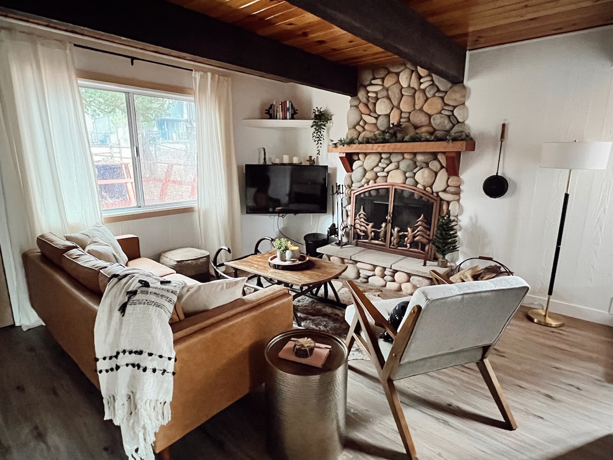 A cozy living area features a stone fireplace, complemented by natural light from the nearby window. Comfortable seating includes a leather sofa and modern armchair, with a wooden coffee table at the center. Decor accents include a light-colored throw and potted plants.