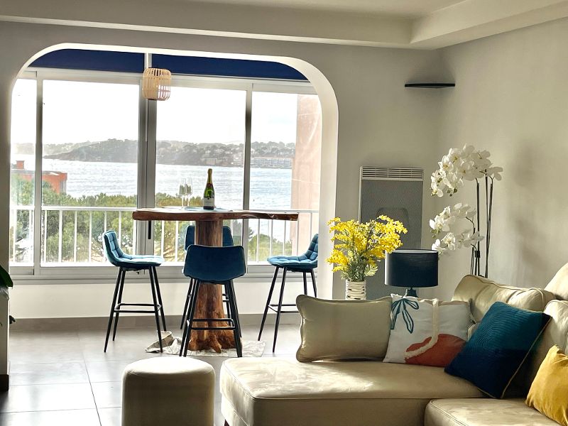 A cozy living area showcases a light-colored sofa and a rustic wooden dining table surrounded by blue bar stools. A vase of yellow flowers and a bottle of champagne sit atop the table. Large windows offer a view of the water and natural light fills the space.