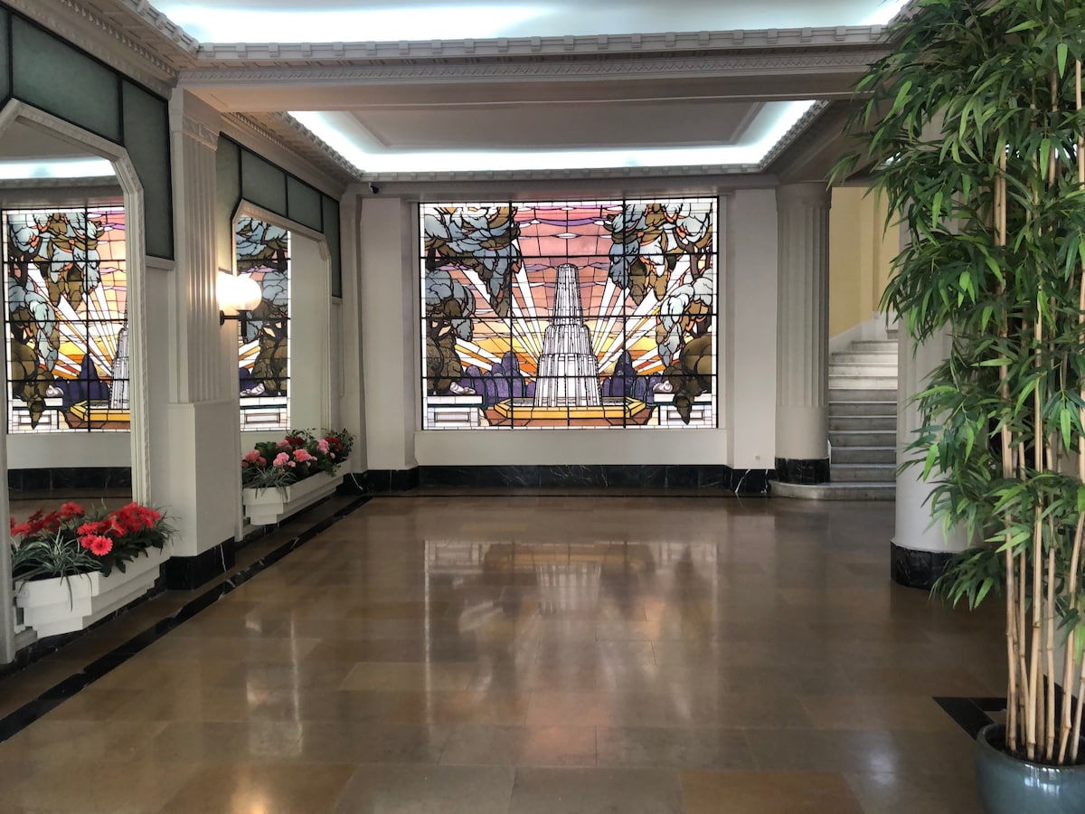 A spacious lobby area features elegant stained glass windows depicting a tranquil scene, allowing natural light to enhance the interior. Potted plants and floral displays line the walls, and polished floors reflect the artistic details and architectural elements present in the space.