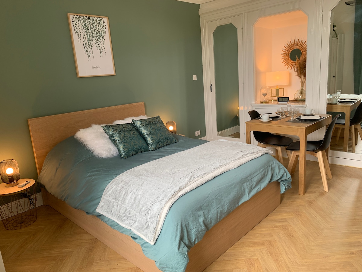 A well-appointed sleeping area features a spacious bed with a soft blue-green cover and decorative pillows. A wooden dining table is set for two, complementing the neutral decor. A reflective wardrobe and wall art add to the overall aesthetic, bathed in soft, natural light.