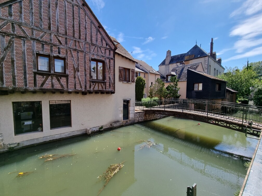 A charming view showcases a traditional building featuring half-timbered architecture beside a calm green pond. A small bridge crosses the water, connecting the buildings in the background. Lush greenery surrounds the area, creating a tranquil outdoor atmosphere.